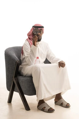 أدوات بصرية للتدقيق والتركيز، مفهوم الفحص والاستكشاف، توضيح الصورة والرؤية عن قرب، بورتريه لرجل عربي خليجي سعودي يرتدي الثوب التقليدي و الشماغ يجلس على كرسي مريح يحمل بيده عدسة مكبرة ينظر من خلالها بإيماءات التركيز والتمعن، خلفية بيضاء