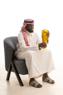 إيماءات وجه تدل على الشعور بالفخر والسعادة بالفوز، تشجيع الفرق والمنتخبات والاندية, بورتريه لرجل عربي خليجي سعودي يرتدي الثوب التقليدي والشماغ يجلس على كرسي يحمل بيديه كأس البطولة ، خلفية بيضاء