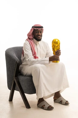 Encouraging teams, national teams, and clubs, a portrait of a Gulf Arab Saudi man wearing traditional attire and a shemagh sitting on a chair holding a championship trophy in his hands, facial expressions indicating feelings of pride and happiness from winning, white background.