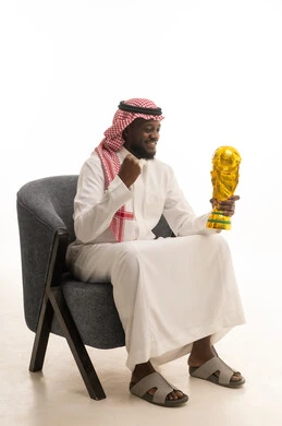 التتويج بالبطولة بعد النجاح، مفهوم الفوز والانتصار، ايماءات السعادة والسرور، بورتريه لشاب عربي خليجي سعودي يرتدي الثوب التقليدي والشماغ يجلس على كرسي ويحمل بيده كأس الفوز، خلفية بيضاء