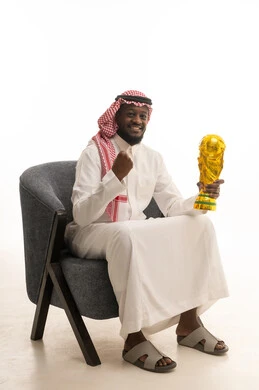 مفهوم الفوز والانتصار، التتويج بالبطولة بعد النجاح، ايماءات السعادة والسرور، بورتريه لشاب عربي خليجي سعودي يرتدي الثوب التقليدي والشماغ يجلس على كرسي ويحمل بيده كأس الفوز، خلفية بيضاء