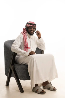 استخدام تقنية الهيدفون، برامج التحدث عن بعد, بورتريه لرجل عربي خليجي سعودي يرتدي الثوب التقليدي و الشماغ و سماعات الرأس يجلس على كرسي يقوم بالرد على اسئلة العملاء بإيماءات البشاشة , العمل في قسم الدعم الفني, خلفية بيضاء