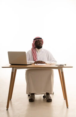 Looking at something, medicine and healthcare, recording important data and notes, a portrait of an Arab Gulf Saudi doctor wearing traditional attire, a ghutrah, and a white coat sitting behind a table in front of a laptop with a stethoscope beside him, white background. Looking at something, medicine and healthcare, recording important data and notes, a portrait of an Arab Gulf Saudi doctor wearing traditional attire, a ghutrah, and a white coat sitting behind a table in front of a laptop with a stethoscope beside him, white background.