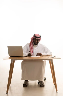 الطب والرعاية الصحية، تسجيل البيانات والملاحظات المهمة، بورتريه لطبيب عربي خليجي سعودي يرتدي الثوب التقليدي والشماغ والمعطف الأبيض يجلس خلف الطاولة امام الحاسب المحمول بجانبة السماعات الطبية ، خلفية بيضاء