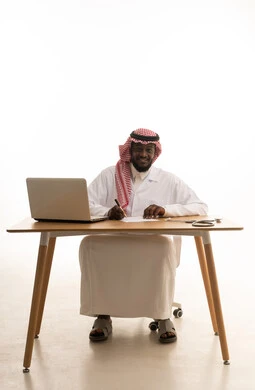 تسجيل البيانات والملاحظات المهمة، الطب والرعاية الصحية، بورتريه لطبيب عربي خليجي سعودي يرتدي الثوب التقليدي والشماغ والمعطف الأبيض يجلس خلف الطاولة امام الحاسب المحمول بجانبة السماعات الطبية ، خلفية بيضاء