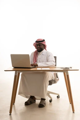 Gestures of happiness and joy, integrating modern technology in the field of medical work, recording and documenting patient information and files via a laptop, a portrait of an Arab Gulf Saudi doctor wearing traditional attire, a shemagh, and a white coat sitting behind a table in front of a laptop with medical headphones beside him, white background.