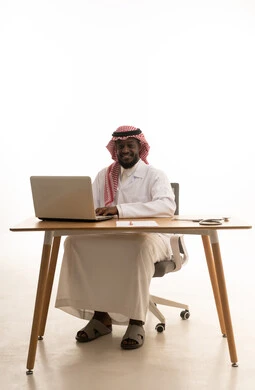 Integrating modern technology in the field of medical work, recording and documenting patient information and files via a laptop, a portrait of an Arab Gulf Saudi doctor wearing traditional attire, a headscarf, and a white coat sitting behind a table in front of a laptop with medical headphones beside him, gestures of happiness and joy, white background.