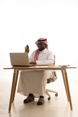 تسجيل وتدوين معلومات وملفات المرضى عبر الابتوب, دمج التقنية الحديثة في مجال العمل الطبي, بورتريه لطبيب عربي خليجي سعودي يرتدي الثوب التقليدي والشماغ والمعطف الأبيض يجلس خلف الطاولة امام الحاسب المحمول بجانبة السماعات الطبية , اجراء مكالمة فيديو، رفع كف اليد بتعابير السلام و التحية ، خلفية بيضاء