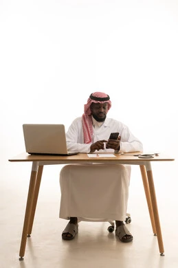 تقديم الخدمات والرعاية الصحية عبر الجوال، استخدام الأجهزة التقنية الحديثة، العمل في قطاع الصحة والرعاية، البرامج والتطبيقات الطبية، بورتريه لطبيب عربي خليجي سعودي يرتدي الثوب التقليدي والشماغ والمعطف الأبيض يجلس خلف الطاولة امام الحاسب المحمول بجانبة السماعات الطبية، خلفية بيضاء