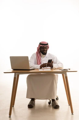 استخدام الأجهزة التقنية الحديثة، العمل في قطاع الصحة والرعاية، البرامج والتطبيقات الطبية، بورتريه لطبيب عربي خليجي سعودي يرتدي الثوب التقليدي والشماغ والمعطف الأبيض يجلس خلف الطاولة امام الحاسب المحمول بجانبة السماعات الطبية، تقديم الخدمات والرعاية الصحية عبر الجوال، خلفية بيضاء