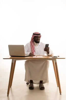 البرامج والتطبيقات الطبية، العمل في قطاع الصحة والرعاية، استخدام الأجهزة التقنية الحديثة، بورتريه لطبيب عربي خليجي سعودي يرتدي الثوب التقليدي والشماغ والمعطف الأبيض يجلس خلف الطاولة امام الحاسب المحمول بجانبة السماعات الطبية، تقديم الخدمات والرعاية الصحية عبر الجوال، خلفية بيضاء