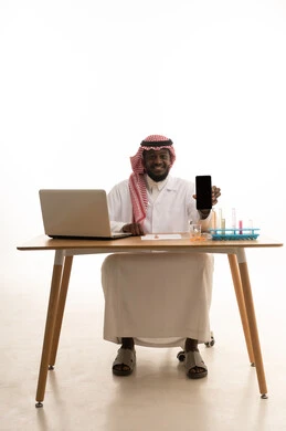The concept of biochemistry, displaying a black screen on a mobile phone, sitting in a chemistry lab, a portrait of an Arab Gulf Saudi doctor wearing a white lab coat and a ghutrah sitting behind a table in front of a laptop, using test tubes to conduct experiments in the lab, white background.