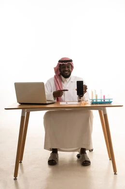 A black screen displayed on a mobile phone, sitting in a chemistry lab, a portrait of an Arab Gulf Saudi doctor wearing a white lab coat and a ghutrah sitting behind a table in front of a laptop, using test tubes to conduct experiments in the lab, the concept of biochemistry, white background.