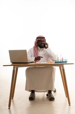 Making a phone call via a mobile phone, sitting in a chemistry lab, a portrait of an Arab Gulf Saudi doctor wearing a white lab coat and a ghutrah sitting behind a table in front of a laptop, using test tubes to conduct experiments in the lab, the concept of biochemistry, white background.