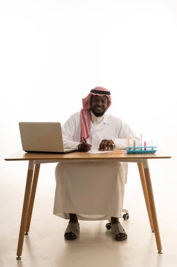 Recording notes and results, sitting in a chemistry lab, a portrait of an Arab Gulf Saudi doctor wearing a white lab coat and a ghutrah sitting behind a table in front of a laptop, using test tubes to conduct experiments in the lab, chemical reactions, the concept of biochemistry, white background.