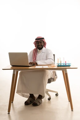 الجلوس في معمل الكيمياء، بورتريه لطبيب عربي خليجي سعودي يرتدي المعطف الطبي الابيض و الشماغ يجلس خلف الطاولة امام الحاسب المحمول , استخدام انابيب الاختبار لاجراء التجارب في المختبر ، التفاعلات الكيميائية ،النقر على لوحة المفاتيح ، مفهوم الكيمياء الحيوية، خلفية بيضاء