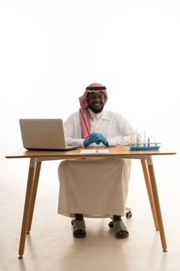 النظر الى الكاميرا بايماءات السعادة و السرور، اكتساب المعرفة والخبرة، التطوير والبحث وإجراء التفاعلات الكيميائية، الدراسات والتجارب في بيئة عملية متطورة, بورتريه لرجل عربي خليجي سعودي يرتدي الزي التقليدي و نظارة الوقاية يجلس امام الحاسوب المحمول، خلفية بيضاء