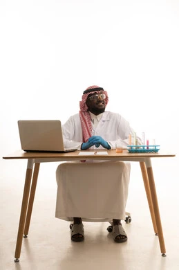 التطوير والبحث وإجراء التفاعلات الكيميائية, اكتساب المعرفة والخبرة، الدراسات والتجارب في بيئة عملية متطورة, بورتريه لرجل عربي خليجي سعودي يرتدي الزي التقليدي و نظارة الوقاية يجلس امام الحاسوب المحمول, النظر الى شيء ما بايماءات السعادة و السرور، خلفية بيضاء