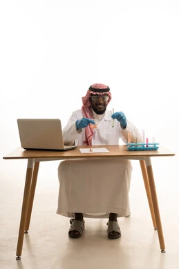 Studying and implementing chemical reactions, using test tubes to conduct experiments and scientific research, research and development, and the advancement of biotechnology, a portrait of a Saudi Arabian Gulf Arab man wearing traditional thobe and shemagh sitting behind a table in front of a laptop examining analytical samples, white background.
