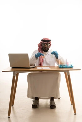 القيام بالتفاعلات الكيميائية وتحليلها، استخدام انابيب الاختبار لإجراء التجارب والبحوث العلمية، البحث والتطوير وتقدم التكنولوجيا الحيوية، بورتريه لرجل عربي خليجي سعودي يتدي الثوب التقليدي و الشماغ  يجلس خلف الطاولة امام الحاسب المحمول يقوم بفحص العينات التحليلية ، خلفية بيضاء