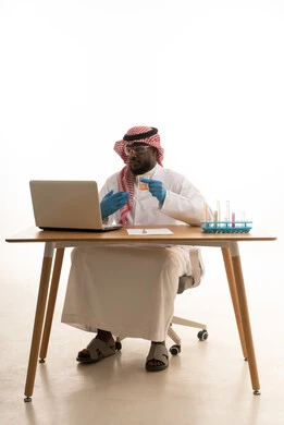 Research and development and the advancement of biotechnology, conducting studies and chemical reactions, using test tubes to carry out experiments and scientific research, a portrait of an Arab Gulf Saudi man wearing traditional attire and a ghutrah sitting behind a table in front of a laptop examining analytical samples, white background.