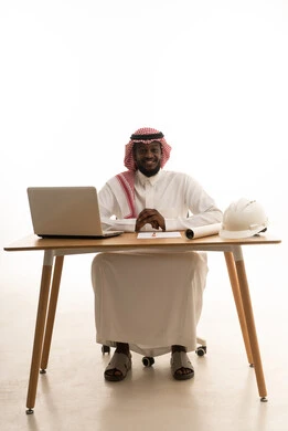 النظر الى الكاميرا بايماءات السعادة و السرور، استخدام جهاز تقني حديث متطور، وظيفة ومهنة سعودية, بورتريه لرجل عربي خليجي سعودي يرتدي الثوب التقليدي و الشماغ يجلس أمام طاولة خشبية و الحاسب المحمول، تدوين الأفكار وتسجيل المعلومات على الأوراق , خلفية بيضاء