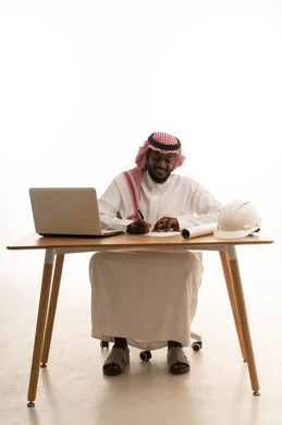 A Saudi job and profession, using a modern advanced technical device, a portrait of a Gulf Arab Saudi man wearing traditional attire and a shemagh sitting in front of a wooden table with a laptop, jotting down ideas and recording information on papers, white background.