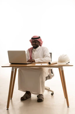 استخدام الأجهزة والتقنيات الحديثة، اجراء مكالمات الفيديو، بورتريه لرجل عربي خليجي سعودي يرتدي الثوب التقليدي و الشماغ يجلس خلف الطاولة يستخدم جهاز الحاسوب المحمول، انجاز مهام العمل عن بعد، الدردشة والتواصل مع الاصدقاء، تصفح الانترنت ومواقع التواصل الاجتماعي، خلفية بيضاء