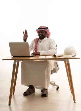 Raising the hand with expressions of peace and greeting, making video calls, using modern technologies in administrative work, a portrait of a Saudi Arabian Gulf man wearing traditional attire and a ghutrah sitting behind a table using a laptop, integrating advanced technology into daily life, white background.