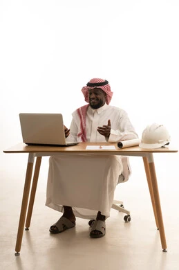 انجاز مهام العمل عن بعد،اجراء مكالمات الفيديو، استخدم التقنيات الحديثة في الأعمال الإدارية ، بورتريه لرجل عربي خليجي سعودي يرتدي الثوب التقليدي و الشماغ يجلس خلف الطاولة يقوم باستخدام الحاسب المحمول ، دمج التكنولوجيا المتطوره فى الحياه اليوميه، خلفية بيضاء