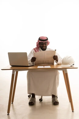 دمج التكنولوجيا بالعمل، تدقيق ومراجعة المخططات الإنشائية، العمل في القطاع الهندسي، إدارة المشاريع الهندسية، ايماءات السرور والسعادة، بورتريه لمهندس عربي خليجي سعودي يرتدي الثوب التقليدي و الشماغ يجلس على الطاولة  امام الحاسب المحمول يقوم بدراسة أساسيات المشروع، خلفية بيضاء