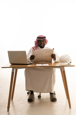 Reviewing and auditing structural plans, working in the engineering sector, managing engineering projects, gestures of joy and happiness, integrating technology into work, a portrait of a Saudi Gulf Arab engineer wearing traditional attire and a shemagh sitting at a table in front of a laptop studying the fundamentals of the project, white background.