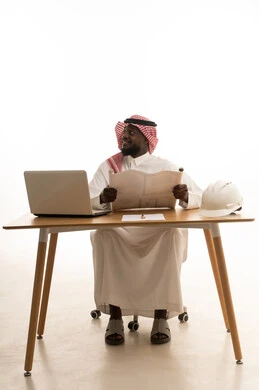 Working in the engineering sector, looking at something, reviewing and auditing construction plans, managing engineering projects, integrating technology into work, a portrait of an Arab Gulf Saudi engineer wearing traditional attire and a ghutrah sitting at a table in front of a laptop studying the fundamentals of the project, white background.