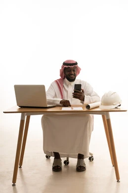 تصفح مواقع التواصل الالكتروني عبر الجوال، بورتريه لمهندس عربي خليجي سعودي يرتدي الثوب الابيض و الشماغ يجلس خلف الطاولة امام الحاسب المحمول بجانبة خوذة الحماية و المخطط الهندسي ، ايماءات السعادة و السرور، خلفية بيضاء