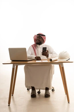 النظر الي شيء ما ، تصفح مواقع التواصل الالكتروني عبر الجوال، بورتريه لمهندس عربي خليجي سعودي يرتدي الثوب الابيض و الشماغ يجلس خلف الطاولة امام الحاسب المحمول بجانبة خوذة الحماية و المخطط الهندسي ، ايماءات السعادة و السرور، خلفية بيضاء