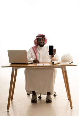 A blank black screen displayed on the mobile, engineering project management, a portrait of an Arab Gulf Saudi engineer wearing a white thobe and a shemagh sitting behind a table in front of a laptop with a safety helmet and engineering plan beside him, gestures of happiness and joy, white background.