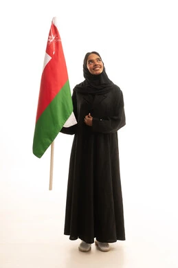 إحياء ذكرى استقلال الدولة 1650 م، الاحتفال باليوم الوطني العماني 18نوفمبر, المشاركة بالاعياد والمناسبات الوطنية، بورتريه لشابة عربية خليجية عمانية ترتدي العباءة والحجاب تحمل بيدها علم سلطنة عمان، النظر بتعابير الفخر والاعتزاز، صورة الجسم بالكامل، خلفية بيضاء