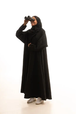 رؤية الأشياء البعيدة، التخطيط المستقبلي للأهداف والطموحات، التفكير والتطلع إلى المستقبل،حب الاستكشاف والمغامرة، بورتريه لشابة عربية خليجية سعودية سمراء ترتدي العباءة والحجاب تمسك منظار ثنائي العينين تنظر إلى شيء ما، الجسم بالطول الكامل، خلفية بيضاء