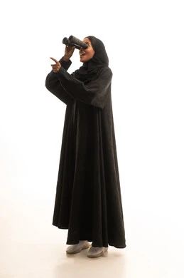 التفكير والتطلع إلى المستقبل، التخطيط المستقبلي للأهداف والطموحات، رؤية الأشياء البعيدة، حب الاستكشاف والمغامرة، بورتريه لشابة عربية خليجية سعودية سمراء ترتدي العباءة والحجاب تمسك منظار ثنائي العينين تنظر إلى شيء ما، الجسم بالطول الكامل، خلفية بيضاء