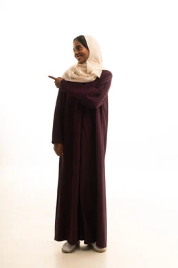 A specific location is identified, a portrait of a smiling Saudi Gulf Arab woman wearing a hijab and abaya, raising her index finger to point at something, with hand gestures indicating this place (here), full-body shot, white background.
