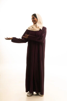 A specific location is identified, a portrait of a smiling Saudi Gulf Arab woman wearing a hijab and abaya, raising her index finger to point at something, with hand gestures indicating this place (here), full-body shot, white background.