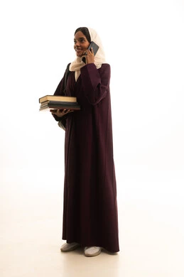 اجراء مكالمة هاتفية ، دمج التقنيات الحديثة بالتعلم والتدريب، التعليم باستخدام التكنولوجيا، بورتريه لطالبة عربية خليجية سعودية ترتدي العباءة و الحجاب  تحمل مجموعة من الكتب تمسك بيدها الهاتف المحمول، خلفية بيضاء