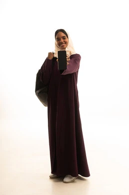 التعليم باستخدام التكنولوجيا،  برامج وتطبيقات التعليم والدراسة اونلاين ، دمج التقنيات الحديثة بالتعلم والتدريب، بورتريه لطالبة عربية خليجية سعودية ترتدي العباءة و الحجاب وحقيبة الظهر تمسك بيدها الهاتف المحمول، خلفية بيضاء