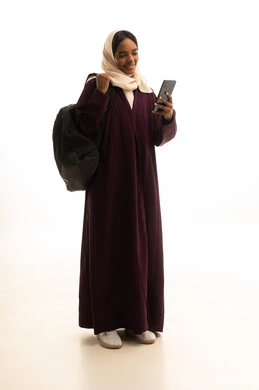 Integrating modern technologies with learning and training, programs and applications for online education and study, education using technology, a portrait of an Arab Gulf Saudi student wearing an abaya and hijab, holding a backpack and a mobile phone in her hand, with a white background.