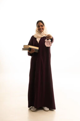 استغلال الوقت في الدراسة والمذاكرة, قدوم وقت الامتحانات والمذاكرات، تطوير المناهج والمواد التعليمية في السعودية، بورتريه لطالبة جامعية عربية خليجية سعودية ترتدي العباءة والحجاب وتحمل بيدها ساعة منبه وكتب, خلفية بيضاء.