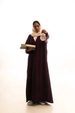 تطوير المناهج والمواد التعليمية في السعودية، قدوم وقت الامتحانات والمذاكرات، استغلال الوقت في الدراسة والمذاكرة, بورتريه لطالبة جامعية عربية خليجية سعودية ترتدي العباءة والحجاب وتحمل بيدها ساعة منبه وكتب, خلفية بيضاء.