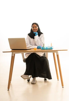 استخدام الأدوات والوسائل التعليمية التجريبية، أساليب التعلم الفعال بالاستفادة من التقنيات والأجهزة التكنولوجية، بورتريه لطالبة عربيه خليجيه سعوديه ترتدي العباءه والحجاب والمعطف الابيض ونظارات الحماية تستخدم جهاز الحاسوب المحمول تجلس امام طاولة الاختبارات وأنابيب العينات والتحاليل الكيميائية، خلفية بيضاء