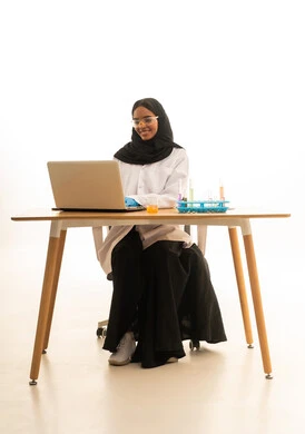 بورتريه لطالبة عربيه خليجيه سعوديه ترتدي العباءه والحجاب والمعطف الابيض ونظارات الحماية تستخدم جهاز الحاسوب المحمول تجلس امام طاولة الاختبارات وأنابيب العينات والتحاليل الكيميائية، أساليب التعلم الفعال بالاستفادة من التقنيات والأجهزة التكنولوجية، استخدام الأدوات والوسائل التعليمية التجريبية،خلفية بيضاء