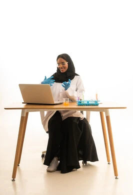 بيئة عملية متطورة ، الاستفادة من التقنيات والأجهزة التكنولوجية، استخدام الأدوات والوسائل التعليمية التجريبية، بورتريه لطالبة عربيه خليجيه سعوديه ترتدي العباءه والحجاب والمعطف الابيض ونظارات الحماية تستخدم جهاز الحاسوب المحمول تجلس امام طاولة الاختبارات وأنابيب العينات والتحاليل الكيميائية، خلفية بيضاء