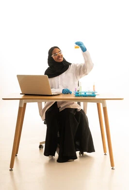 استخدام الأدوات والوسائل التعليمية التجريبية، أساليب التعلم الفعال بالاستفادة من التقنيات والأجهزة التكنولوجية، بورتريه لطالبة عربيه خليجيه سعوديه ترتدي العباءه والحجاب والمعطف الابيض ونظارات الحماية تستخدم جهاز الحاسوب المحمول تجلس امام طاولة الاختبارات وأنابيب العينات والتحاليل الكيميائية، خلفية بيضاء